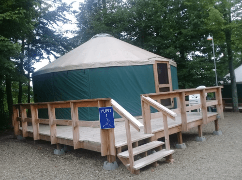 A green yurt on a wooden deck surrounded by trees, with a sign reading "Yurt 1" and steps leading up to it.